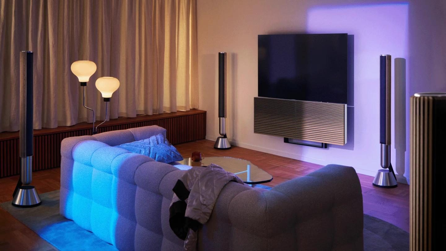 An evening living room lit by floor lamps and ambient lighting, with a wall-mounted television and tall speakers